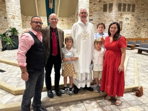 Family photo after Jamie and William's Catholic baptism — Erjon, Blane, Deacon Dan, Jamie, William, Michael, and Sharon standing together in the church.