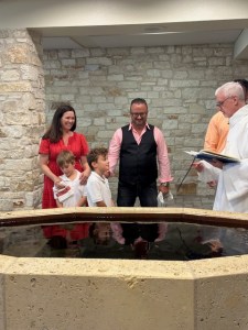 Jamie and William standing at the baptismal font with Sharon and Erjon beside them and Deacon Dan reading from a book.